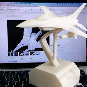 May include: A white 3D printed model of a fighter jet on a stand. The model is displayed on a laptop computer screen with a website open in the background.