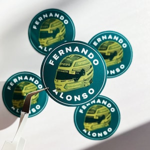 Puede incluir: Cuatro pegatinas redondas de color azul verdoso con el texto "Fernando Alonso" en letras blancas. Cada pegatina presenta un casco de carreras amarillo y negro.