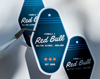 Op de afbeelding: Drie blauwe en witte motel-sleutelhanger stickers met de tekst "Formule 1 Red Bull Milton Keynes, Engeland Est 2005" erop gedrukt.