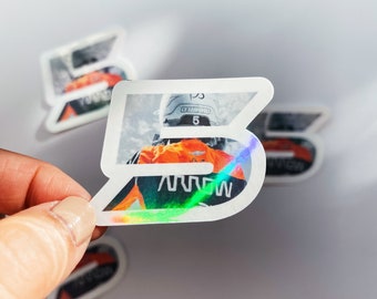 May include: A white sticker with a holographic finish featuring a skier wearing a helmet and a racing suit. The sticker has the number "5" and the word "ARROW" printed on it.