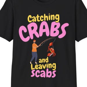 May include: Black t-shirt with a humorous graphic of a person fishing for crabs. The text reads "Catching Crabs and Leaving Scabs".