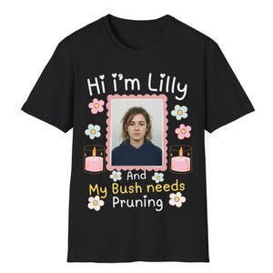 Puede incluir: Una camiseta negra de manga corta con una foto rectangular central de una persona, rodeada de ilustraciones de flores rosas y azules y dos velas rosas. El texto dice: Hi i'm Lilly And My Bush needs Pruning en blanco y amarillo.