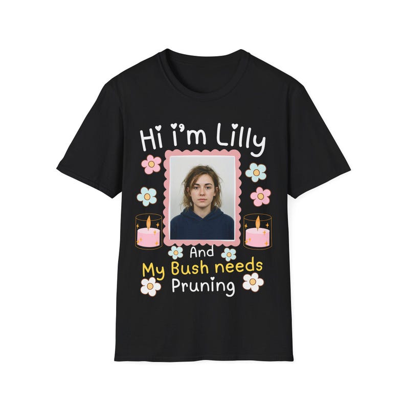 Puede incluir: Una camiseta negra de manga corta con una foto rectangular central de una persona, rodeada de ilustraciones de flores rosas y azules y dos velas rosas. El texto dice: Hi i'm Lilly And My Bush needs Pruning en blanco y amarillo.