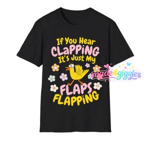 Może przedstawiać: Czarny t-shirt z napisem "If You Hear CLAPPING It's Just My FLAPS FLAPPING" w kolorze r&oacute;żowym i ż&oacute;łtym. Ż&oacute;łta kresk&oacute;wkowa kaczka z trzepoczącymi skrzydłami znajduje się na środku, otoczona kwiatami. Koszulka ma okrągły dekolt i kr&oacute;tkie rękawy.