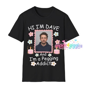 May include: Black t-shirt featuring the text "HI I'M DAVE" above a framed photo of a man. The text "And I'm a Pegging Addict" is below the photo. The design includes pink and blue floral accents and two pink candles.