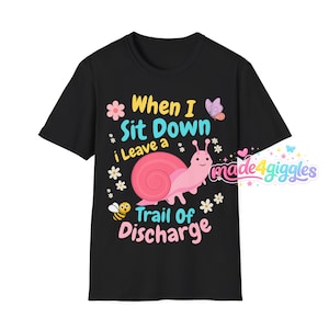 May include: A black short-sleeve t-shirt featuring a pink snail graphic and the text "When I Sit Down i Leave a Trail Of Discharge". The design includes small flowers, a bee, and a butterfly, with the brand name "made4giggles" in the corner.