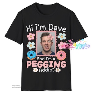 May include: A black t-shirt featuring a central photograph of a person with a pink bow graphic, surrounded by pastel flowers and two cartoon donuts. The text reads Hi i'm Dave and And i'm a PEGGING Addict in white and pink lettering.