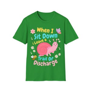 May include: A green short-sleeve t-shirt featuring a pink snail graphic, a small bee, a butterfly, and white flowers. The text reads: When I Sit Down i Leave a Trail Of Discharge. The design uses yellow, light blue, and pink lettering.