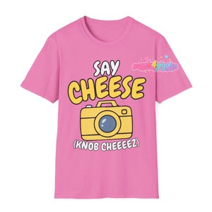 May include: Pink t-shirt with a yellow cartoon camera graphic and the words "SAY CHEESE" above it. Below the camera is the phrase "(KNOB CHEEEZ)". The shirt has short sleeves and a crew neck.