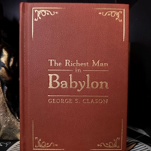 May include: A hardcover book titled "The Richest Man in Babylon" by George S. Clason. The book has a reddish-brown cover with gold lettering and decorative gold accents. The book is a financial literacy guide.