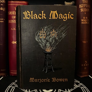 May include: A black book with gold lettering titled "Black Magic" by Marjorie Bowen. The cover features a stylised illustration of a burning brazier with two crowns above it.