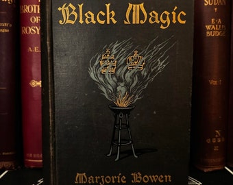May include: A black book with gold lettering titled "Black Magic" by Marjorie Bowen. The cover features a stylised illustration of a burning brazier with two crowns above it.