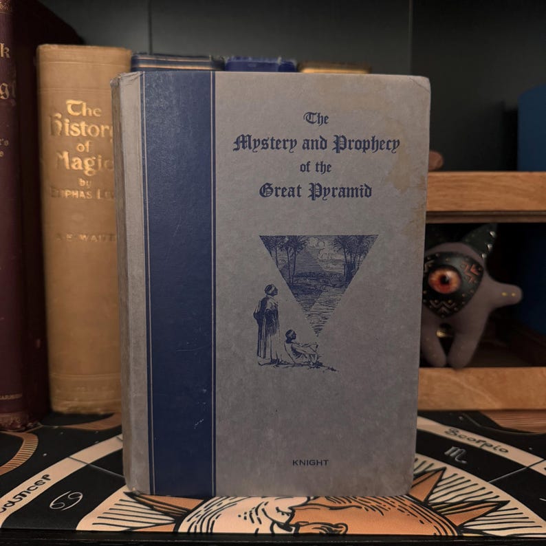 May include: A vintage book titled "The Mystery and Prophecy of the Great Pyramid" with a blue spine and a gray cover. The cover features a black and white illustration of a pyramid with two figures standing in front of it.