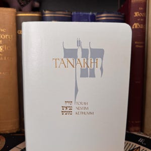 May include: A white book with the word "TANAKH" in gold lettering and a gray graphic. Below the title are Hebrew words and English translations. The book is surrounded by other books.