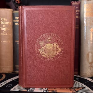 Può includere: Un libro rosso rilegato con un designo in rilievo dorato sulla copertina. Il design presenta un globo con una rosa dei venti e una lettera G al centro. Il libro è intitolato "La Storia della Magia" di Eliphas Levi.