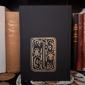 May include: A black book with a gold foil design on the cover. The design is an abstract illustration of figures and shapes. The book is surrounded by other antique books with various colors and titles. The book is standing upright.