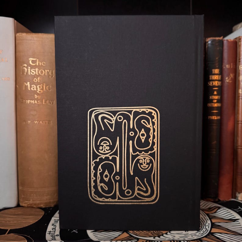 May include: A black book with a gold foil design on the cover. The design is an abstract illustration of figures and shapes. The book is surrounded by other antique books with various colors and titles. The book is standing upright.