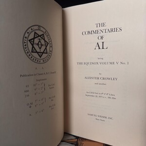 May include: Open book with the title "The Commentaries of AL" by Aleister Crowley. The book's pages are off-white, with text in black. A circular emblem with a star and text is on the left page. The book is part of "The Equinox Volume V No. 1".