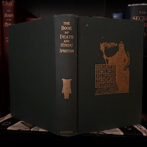 May include: A dark green hardcover book titled "The Book of Death and Hindu Spiritism" with gold lettering. The cover features a gold illustration of a figure holding a torch and a grid with the word "DEATH" spelled out.