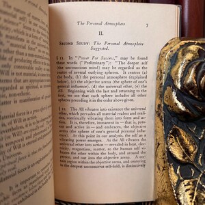 Puede incluir: Libro abierto con texto sobre "La Atm&oacute;sfera Personal". Las p&aacute;ginas est&aacute;n envejecidas, con texto visible y un borde decorativo de color dorado. El texto incluye las palabras "Power For Success" y "universal ether".