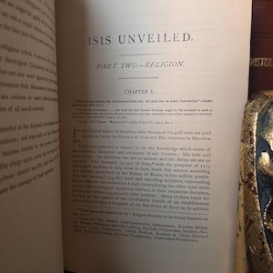 May include: Open book with text titled "ISIS UNVEILED" and "PART TWO-RELIGION." Chapter 1 is visible, with text discussing religion and science. The pages are aged, with visible text and a vintage aesthetic.