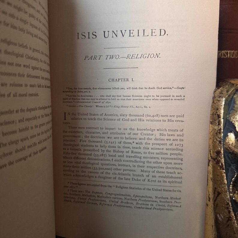 May include: Open book with text titled "ISIS UNVEILED" and "PART TWO-RELIGION." Chapter 1 is visible, with text discussing religion and science. The pages are aged, with visible text and a vintage aesthetic.