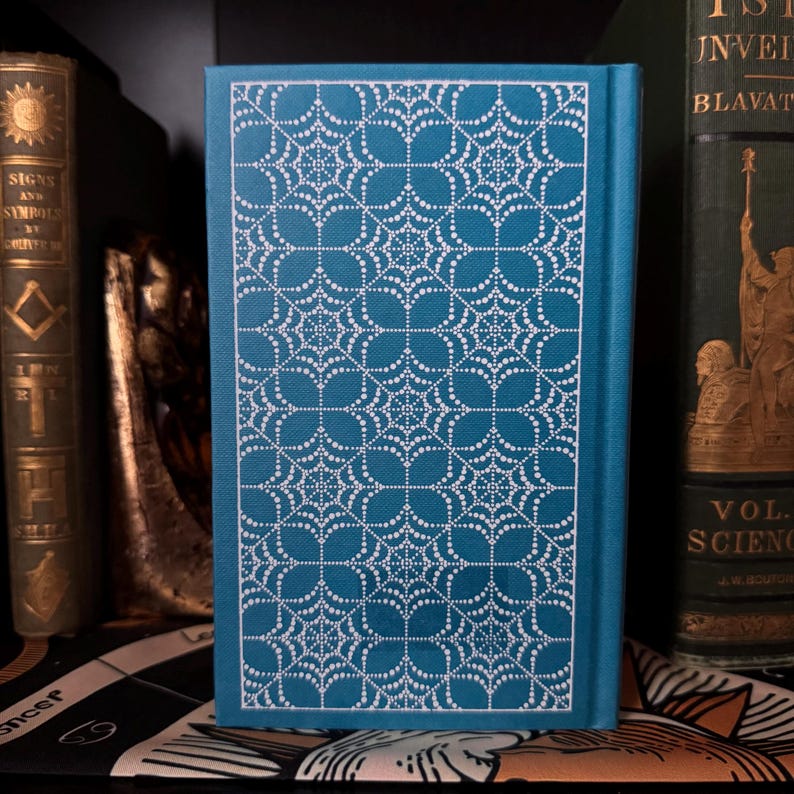 May include: A teal book with a white, intricate, geometric pattern on the cover. The spine is teal. Other antique books are visible in the background, suggesting a collection or library setting.