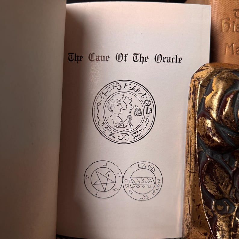 May include: An open book with the title "The Cave Of The Oracle" printed in black. The page features a circular illustration of a figure holding a flower, and two additional circular symbols below. A decorative gold and brown object is visible on the right.
