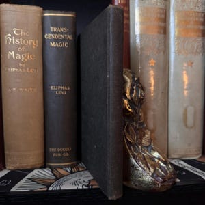 Op de afbeelding: Een verzameling antieke boeken met gouden letters, waaronder "The History of Magic" en "Transcendental Magic." Een decoratieve gouden boekensteun met een sierlijk ontwerp ondersteunt de boeken op een plank.