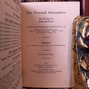 Puede incluir: Libro abierto titulado "The Personal Atmosphere" de Frank Channing Haddock, Ph.D. La portada del libro es de color beige claro con texto negro. El texto incluye "Ten Studies in Poise and Power" y "Twenty-First Edition."