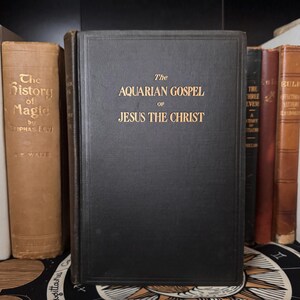 May include: A collection of antique books, including a prominent black volume titled "The Aquarian Gospel of Jesus the Christ" in gold lettering. Other books have brown and red covers. The book spines are visible.