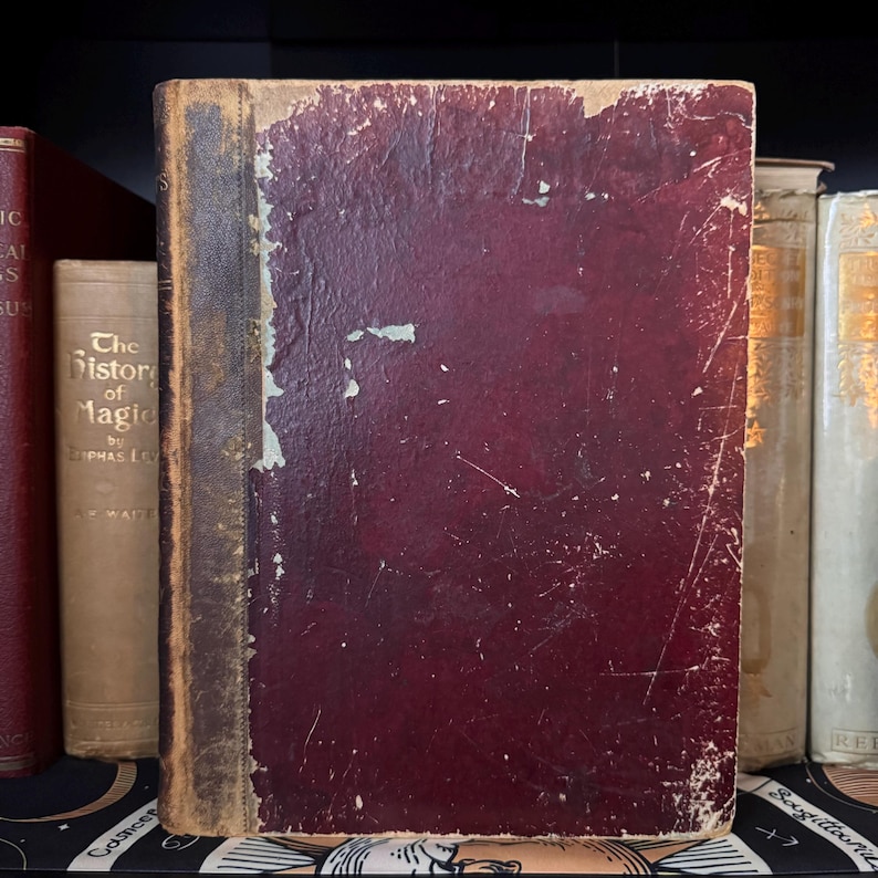 Puede incluir: Un libro antiguo con una cubierta burdeos desgastada y un lomo deteriorado. El libro forma parte de una colecci&oacute;n de libros antiguos, con t&iacute;tulos visibles en los lomos, incluyendo "The History of Magic". La apariencia envejecida del libro sugiere que es un art&iacute;culo de colecci&oacute;n.