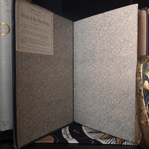 May include: An open antique book with a blue cover and patterned endpapers. The title "Library of Mrs. More Wicks" is visible on a label. The pages are aged, and the book is displayed against a dark background.