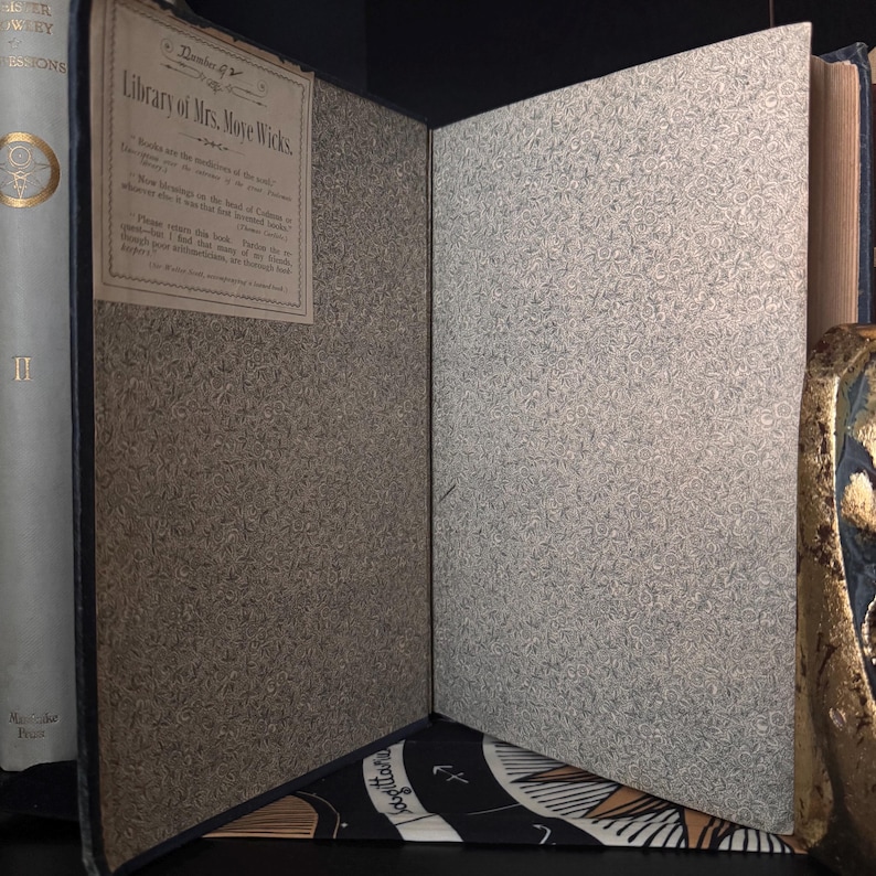 May include: An open antique book with a blue cover and patterned endpapers. The title "Library of Mrs. More Wicks" is visible on a label. The pages are aged, and the book is displayed against a dark background.