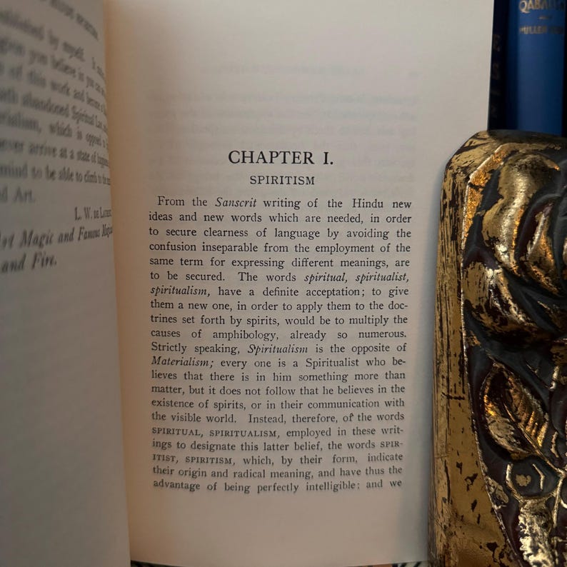 May include: A vintage book with a gold-colored bookmark. The book's title is "Chapter I. Spiritism" and the text discusses the origin of the word "spiritualism" and its meaning.