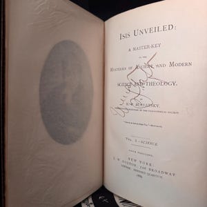 May include: Open book titled "Isis Unveiled: A Master-Key to the Mysteries of Ancient and Modern Science and Theology" by H.P. Blavatsky. The book is open to the title page, with a signature and the publisher's information.