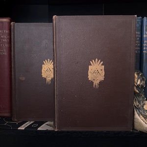 Puede incluir: Dos libros antiguos de cuero marr&oacute;n con s&iacute;mbolos mas&oacute;nicos dorados en relieve. Los libros est&aacute;n de pie sobre una estanter&iacute;a, con otros libros al fondo. Los libros tienen una est&eacute;tica cl&aacute;sica y vintage.
