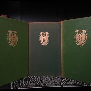 Könnte beinhalten: Drei grüne Hardcover-Bücher mit goldener Beschriftung und einem goldenen Adleremblem auf dem vorderen Umschlag. Die Bücher sind nebeneinander auf einer schwarz-weißen gemusterten Oberfläche angeordnet.