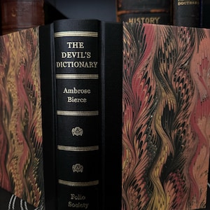 May include: A black leather-bound book with gold lettering titled "The Devil's Dictionary" by Ambrose Bierce. The book is part of the Folio Society collection and has a marbled cover design.
