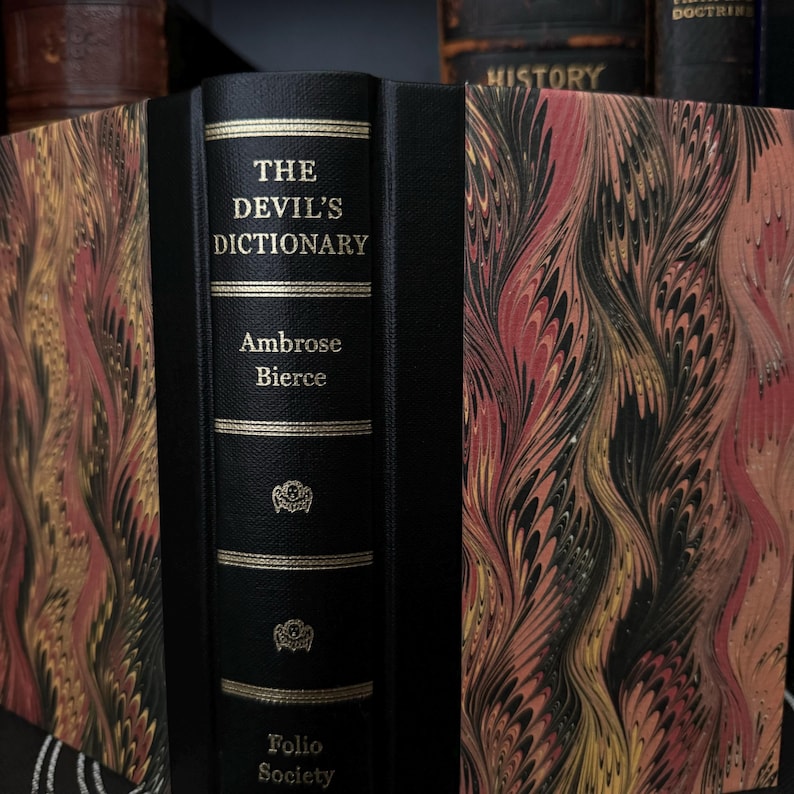 May include: A black leather-bound book with gold lettering titled "The Devil's Dictionary" by Ambrose Bierce. The book is part of the Folio Society collection and has a marbled cover design.
