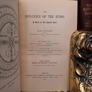 May include: A vintage book titled "The Influence of the Stars: A Book of Old World Lore" by Rosa Baughan. The book cover is cream-colored with black text and is illustrated with a black and white diagram of a circle with numbers and symbols.