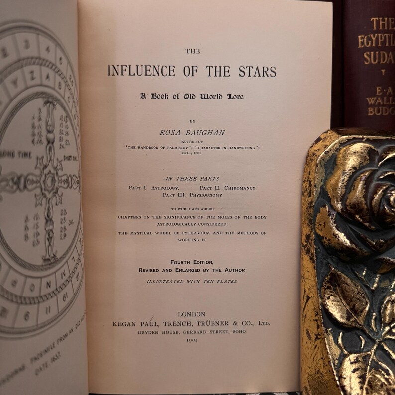 May include: A vintage book titled "The Influence of the Stars: A Book of Old World Lore" by Rosa Baughan. The book cover is cream-colored with black text and is illustrated with a black and white diagram of a circle with numbers and symbols.