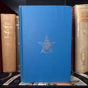 Op de afbeelding: Een blauw hardback boek met een gouden pentagram op de voorkant. Het boek staat in een boekenkast met andere boeken.