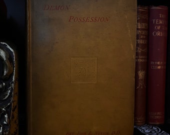 Peut inclure: Un livre vintage intitulé "Possession démoniaque" par Rev. John L. Nevius, D.D. La couverture du livre est de couleur brun foncé avec des lettres dorées et un motif en relief doré.