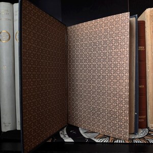 May include: An open book with a dark brown cover and a patterned interior. The pages have a light tan background with a repeating black geometric design. Other books are visible in the background, with white and brown covers.