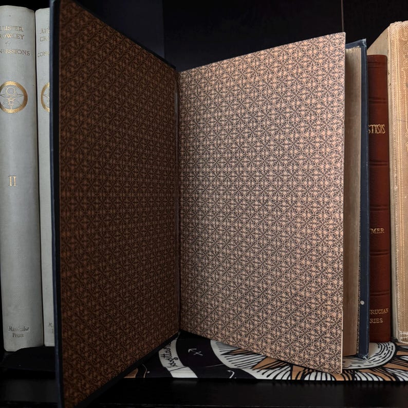May include: An open book with a dark brown cover and a patterned interior. The pages have a light tan background with a repeating black geometric design. Other books are visible in the background, with white and brown covers.