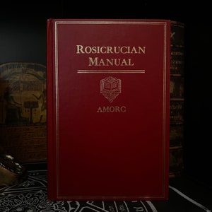以下が含まれることがあります： 金色の文字で「Rosicrucian Manual」と書かれた赤いハードカバーの本。本には金のエンブレムと金のトリムが付いています。AMORCのロゴが下に表示されています。