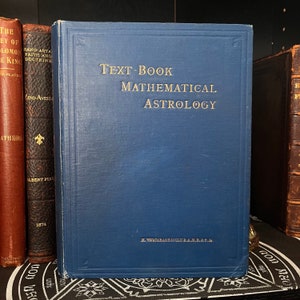 May include: A vintage blue book titled "Text-Book Mathematical Astrology" with gold lettering. The book is standing upright next to other antique books with brown and red covers. The author's name is printed at the bottom.