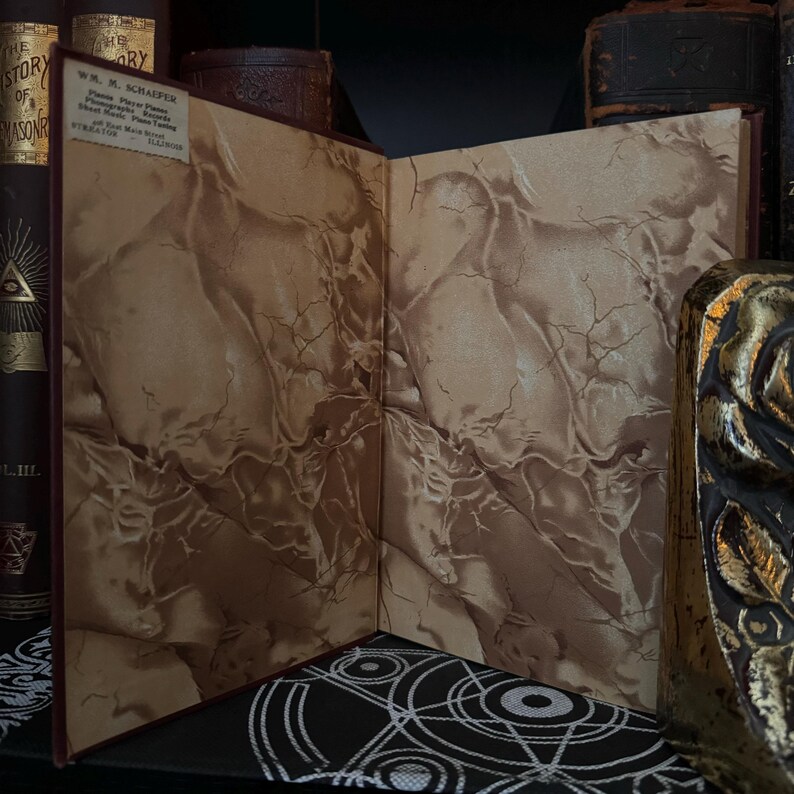 May include: An open book with marbled endpapers. The book is bound in red leather and has a label on the front cover that reads "W.M. Schaffer, Piano, Organ, and Church Music, Publisher, 401 Main Street,  Telephone 1111."