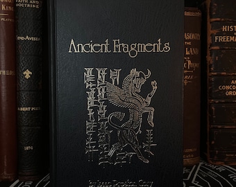 May include: A black hardcover book titled "Ancient Fragments" by Isaac Preston Cory. The book cover features a gold embossed illustration of a winged creature with a bull's head.